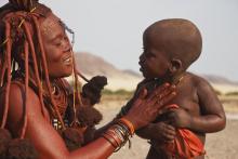 A traditional Himba woman covers her son with a fat and ochre mixture to protect him from sun, Kaokoland, Namibia.